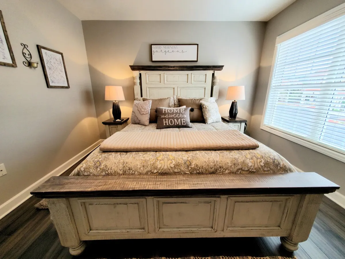 A warmly lit bedroom scene with a bed covered in decorative pillows, nightstands with lamps, framed art on the walls, and a large window with blinds. The ambiance feels cozy and welcoming, ideal for relaxation and reading.