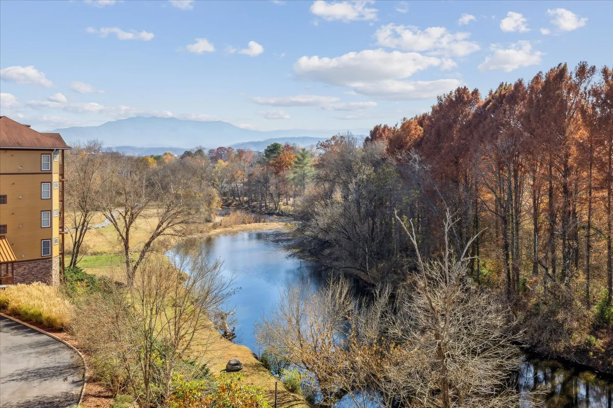 A tranquil riverside scene with mountains in the background, autumn-hued trees lining a peaceful riverbank, and a building structure visible along the water. The atmosphere is serene, suggesting opportunities for nature walks, picnics, and enjoying scenic beauty.