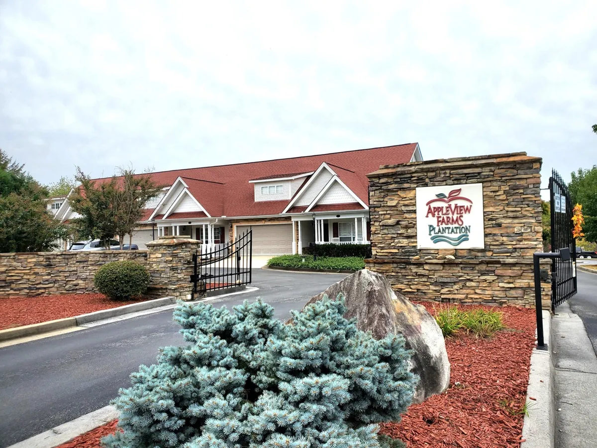 A welcoming exterior view of the resort entrance with a prominent stone sign, landscaped greenery, and a red-roofed building in the background. The scene suggests a community-oriented atmosphere and serves as a point of return after outdoor adventures.