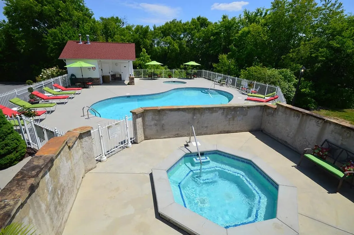 A vibrant exterior shot of the resort's swimming pool area, featuring a sparkling pool, jacuzzi, poolside lounge chairs with green umbrellas, and a pool house in the background. The setting is lively, inviting, and perfect for family fun and socializing.