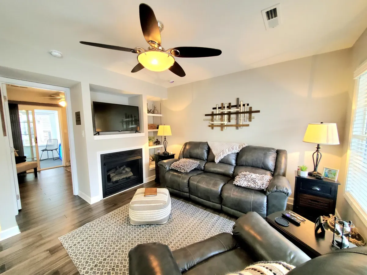 This image features a welcoming living room with a leather sofa facing a fireplace, a TV mounted above, decorative wall art, and warm lighting from table lamps. The layout invites relaxation or social gatherings, highlighting the coziness of the fire and comfortable seating.