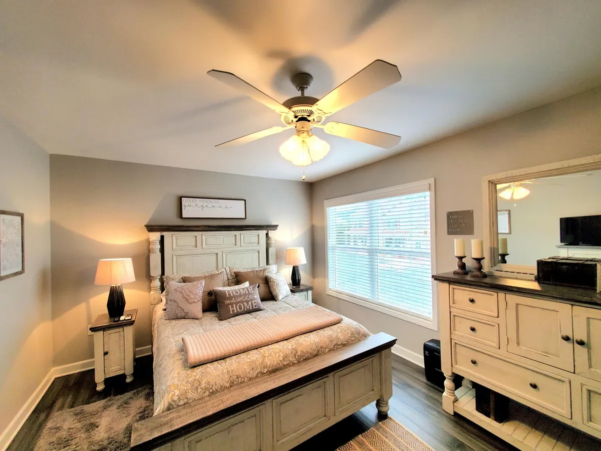 This image features a tranquil bedroom with a bed adorned with decorative pillows, nightstands with lamps on either side, and a dressing table with a mirror. Soft lighting and a neutral color palette create a serene, comfortable atmosphere.
