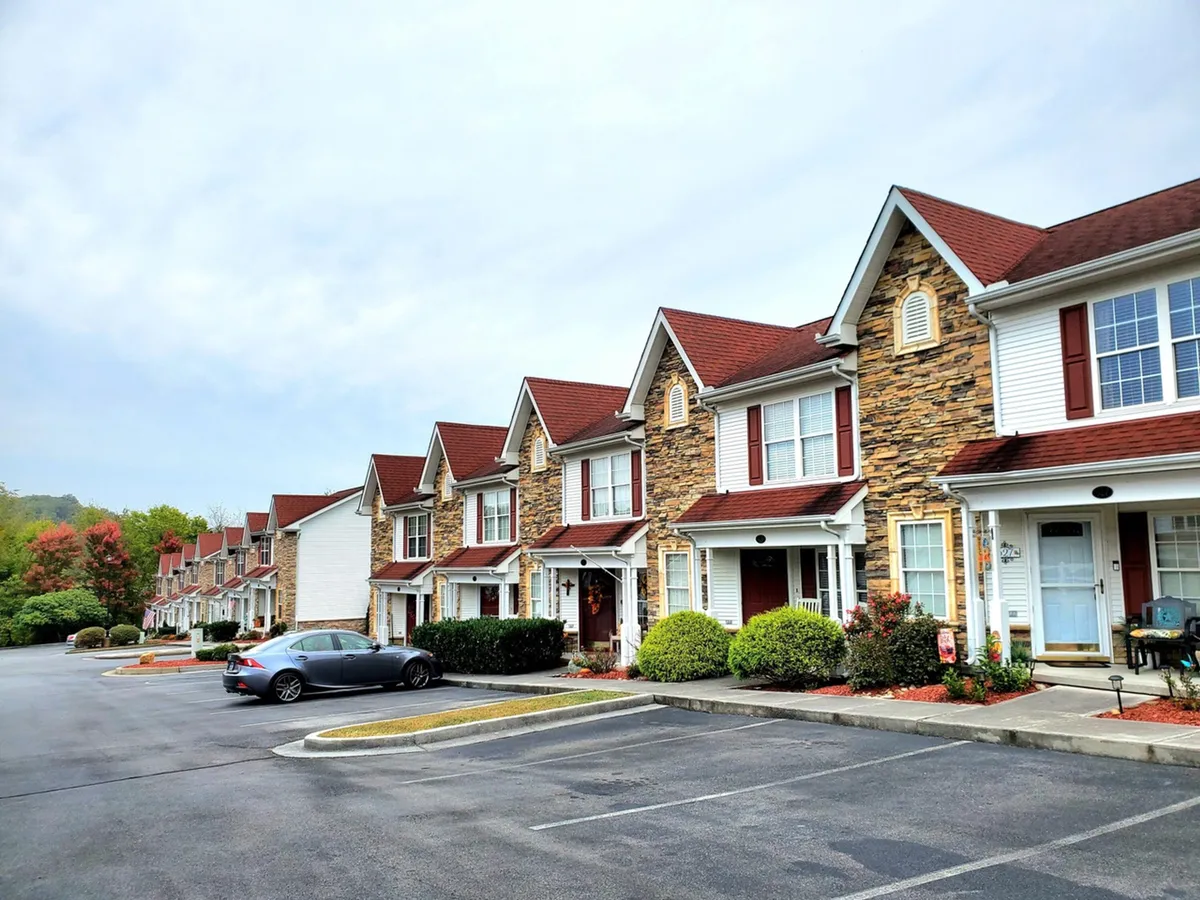 The image showcases a row of attractive townhome-style condos nestled among manicured, landscaped gardens. A parked car hints at easy access, while people are seen walking, socializing, and relaxing outdoors, contributing to a friendly neighborhood atmosphere. Subtle holiday decorations and inviting green spaces evoke a sense of togetherness and celebration.