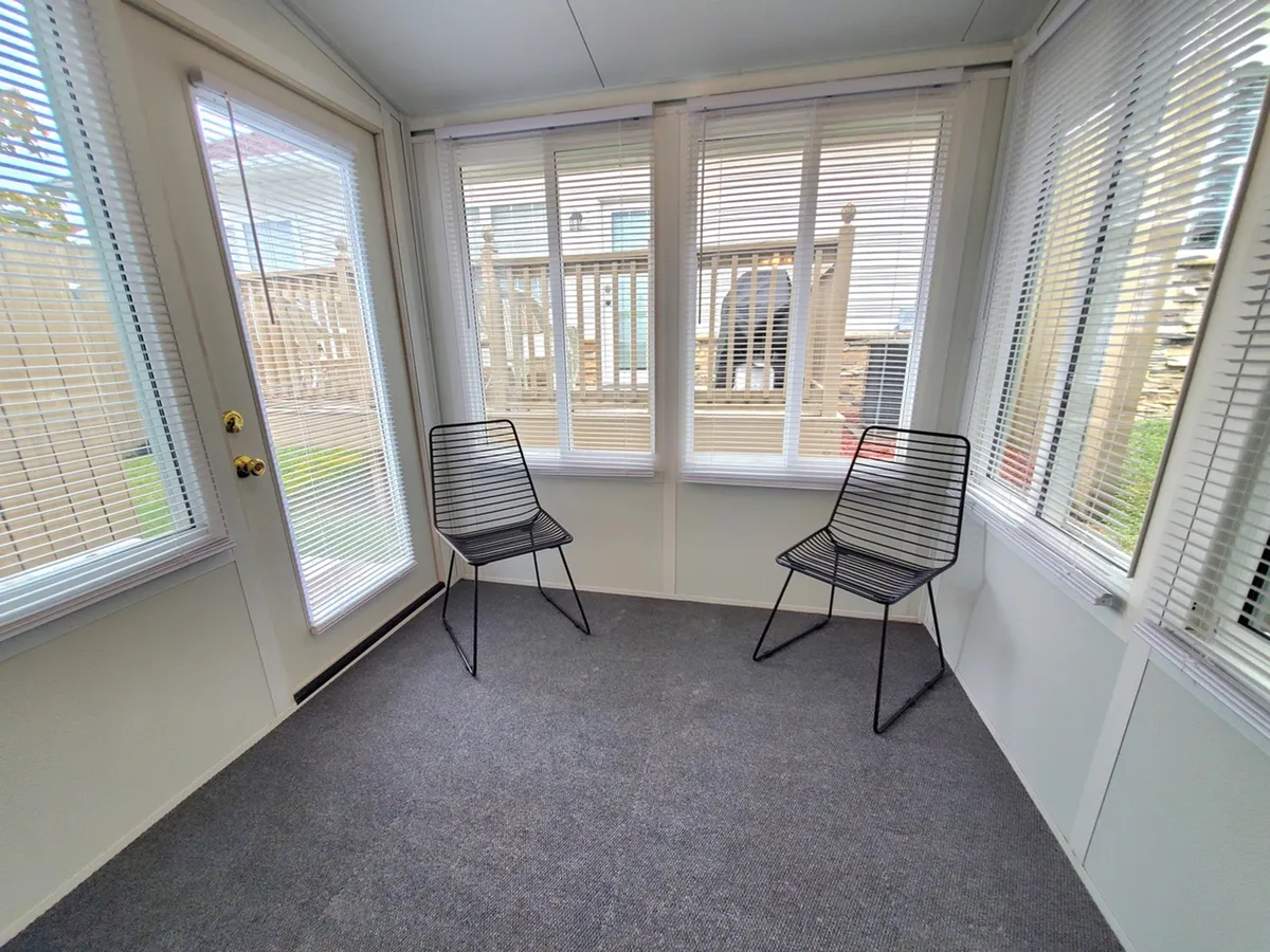 A serene sunroom featuring two modern black wire chairs positioned near large windows with blinds, and a glass door that opens to the outdoors. The space is bathed in natural light, creating an inviting area for morning coffee or evening relaxation.