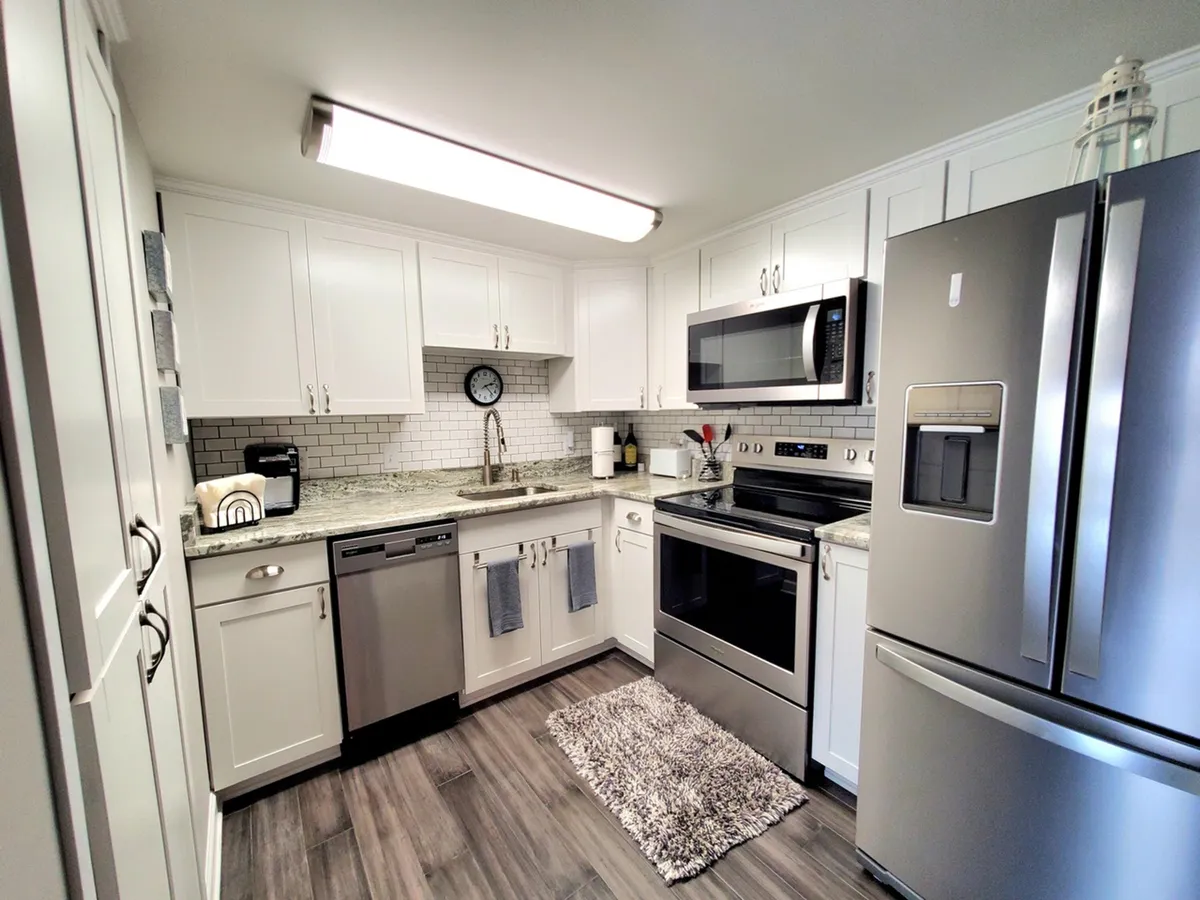 This image features a modern, fully-equipped kitchen with granite countertops, stainless steel appliances, and white cabinetry. The space is clean and functional, with a clock on the wall and ample surfaces for cooking and baking.