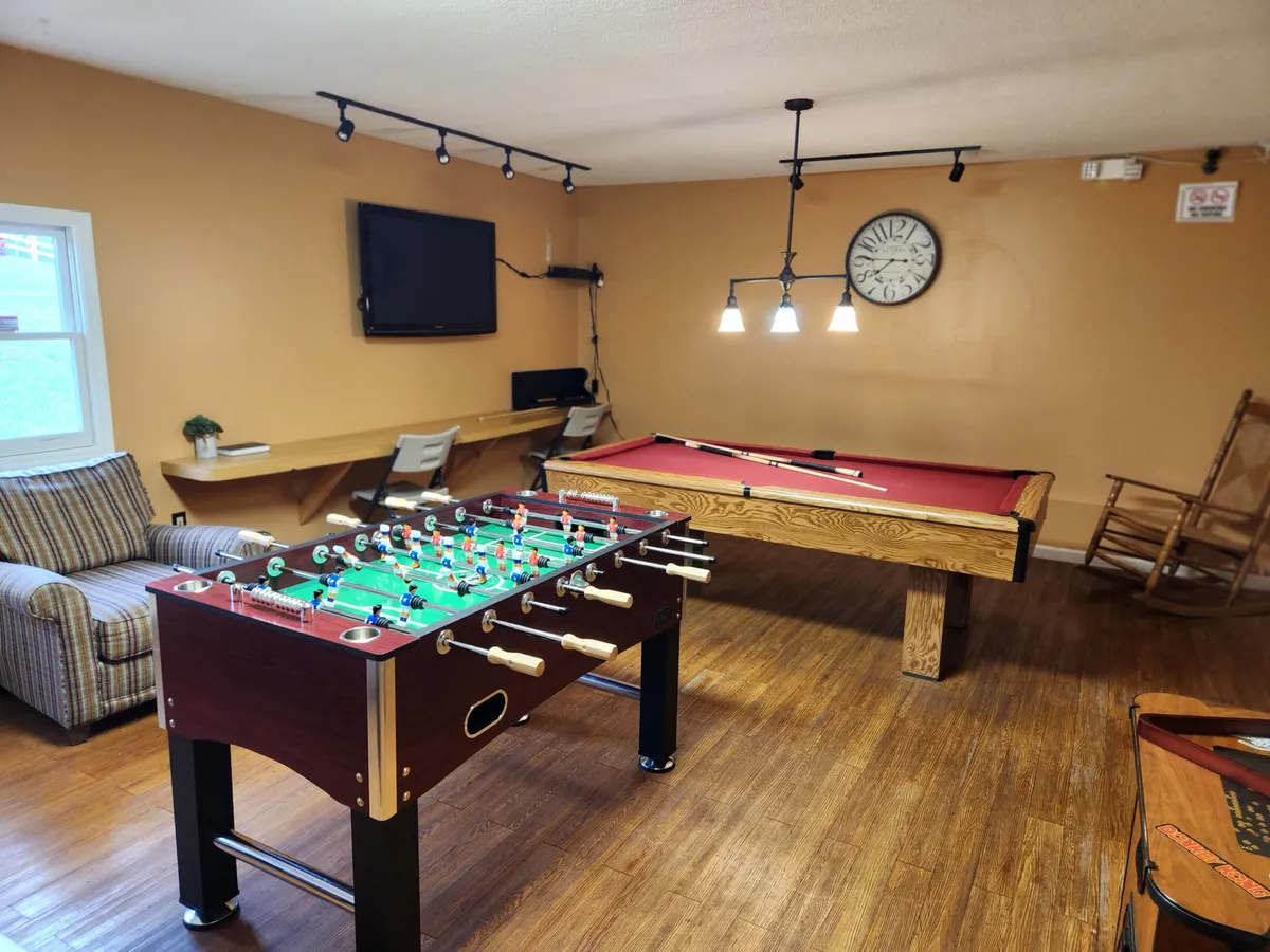 A vibrant interior game room featuring a foosball table, a pool table, a cozy rocking chair, and a television, all set in a playful yet relaxed atmosphere. The space is designed for friendly competition and social interaction, with ample room for both adults and kids to have fun.